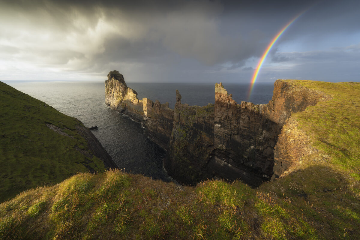 Tory Island cliffs in County Donegal, Ireland, with dramatic sea stacks and a rainbow forming as a rain squall moves in over the Atlantic Ocean under stormy skies.