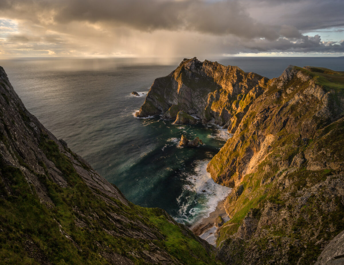 Dramatic sea cliffs in County Donegal, Ireland, at sunset, with a remote cove, turquoise water, and rain showers over the Atlantic Ocean under moody skies.