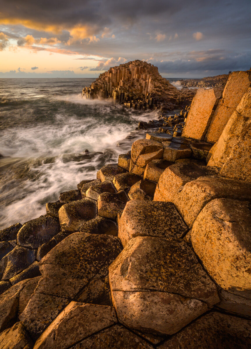 Giant’s Causeway in Northern Ireland at sunset, with hexagonal basalt columns lit by warm golden light and waves crashing along the rugged Atlantic coastline.