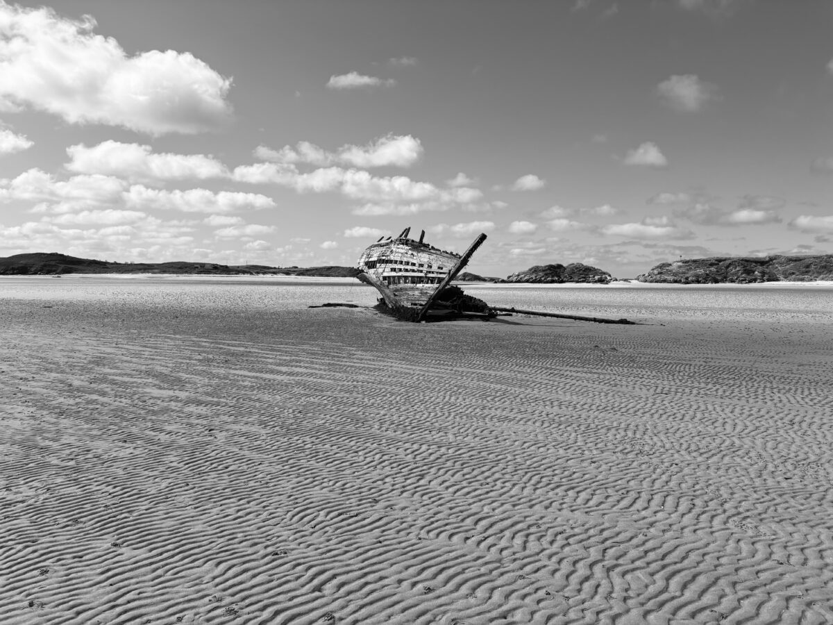 Black and white photograph of a shipwreck on a sandy tidal flat in County Donegal, Ireland, with rippled sand patterns and distant low hills under scattered clouds.