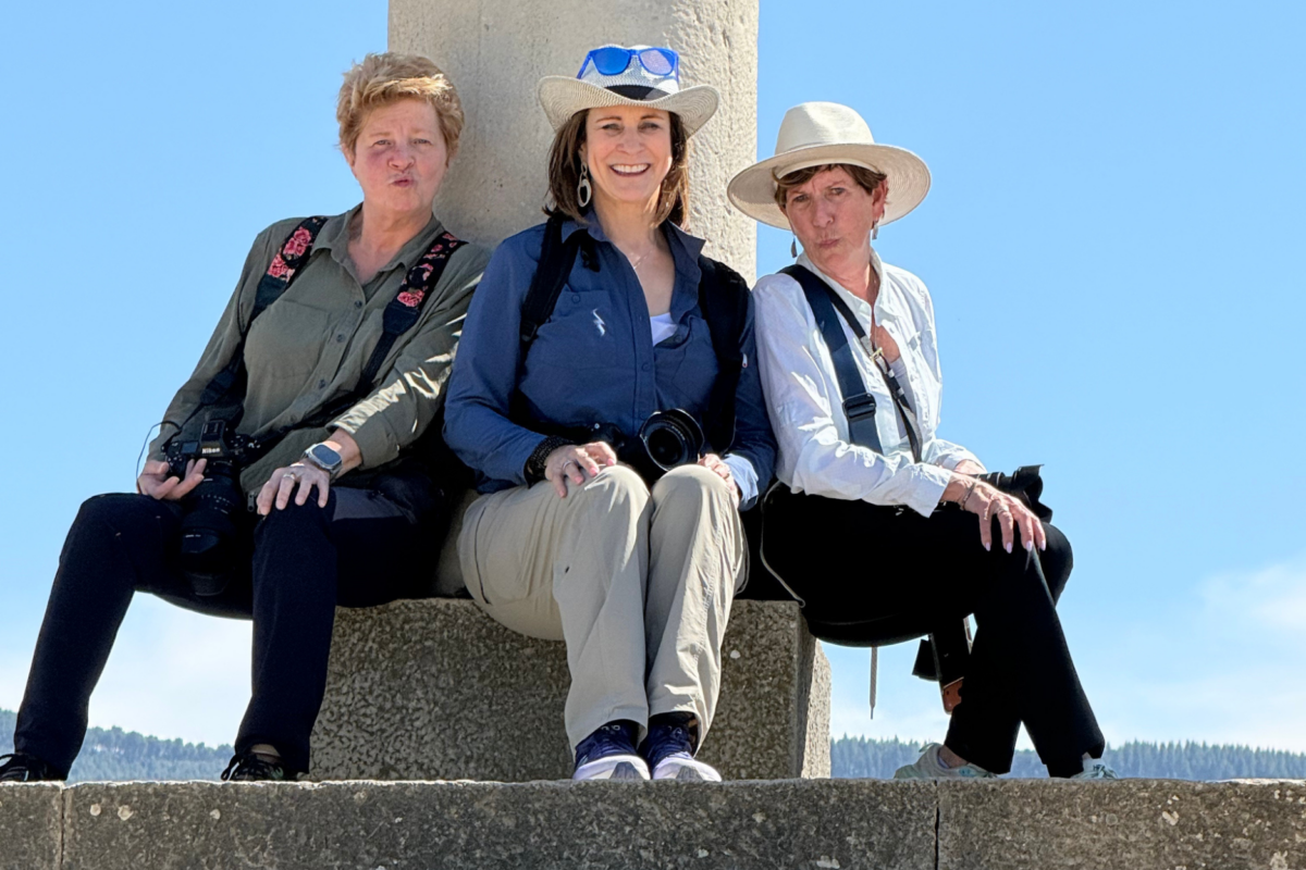 Three female photographers sit together on stone steps beneath a monument, smiling and posing with cameras during a photography outing.