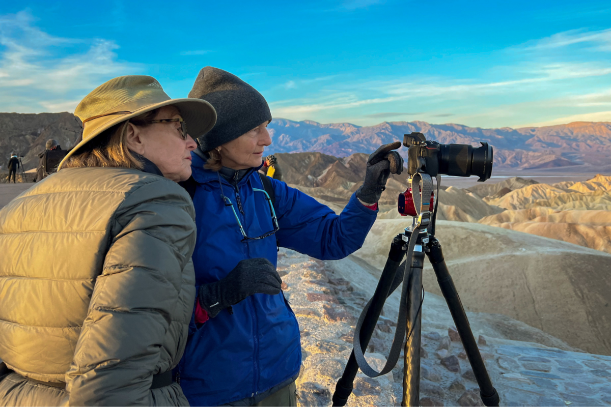 Two female photographers review an image on a camera mounted to a tripod while standing at a scenic overlook during golden-hour light.