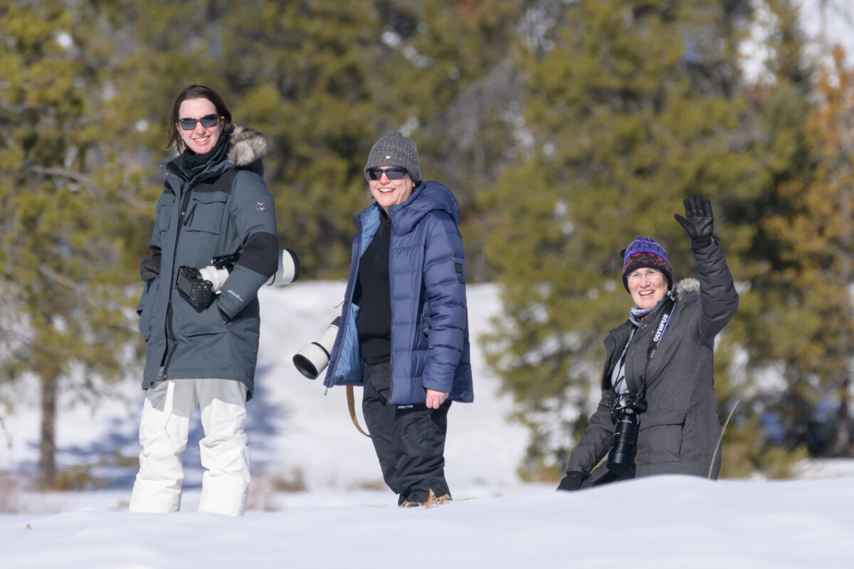 Three photographers stand together in a snowy landscape, bundled against the cold and carrying cameras with telephoto lenses. One waves toward the camera while the others smile.