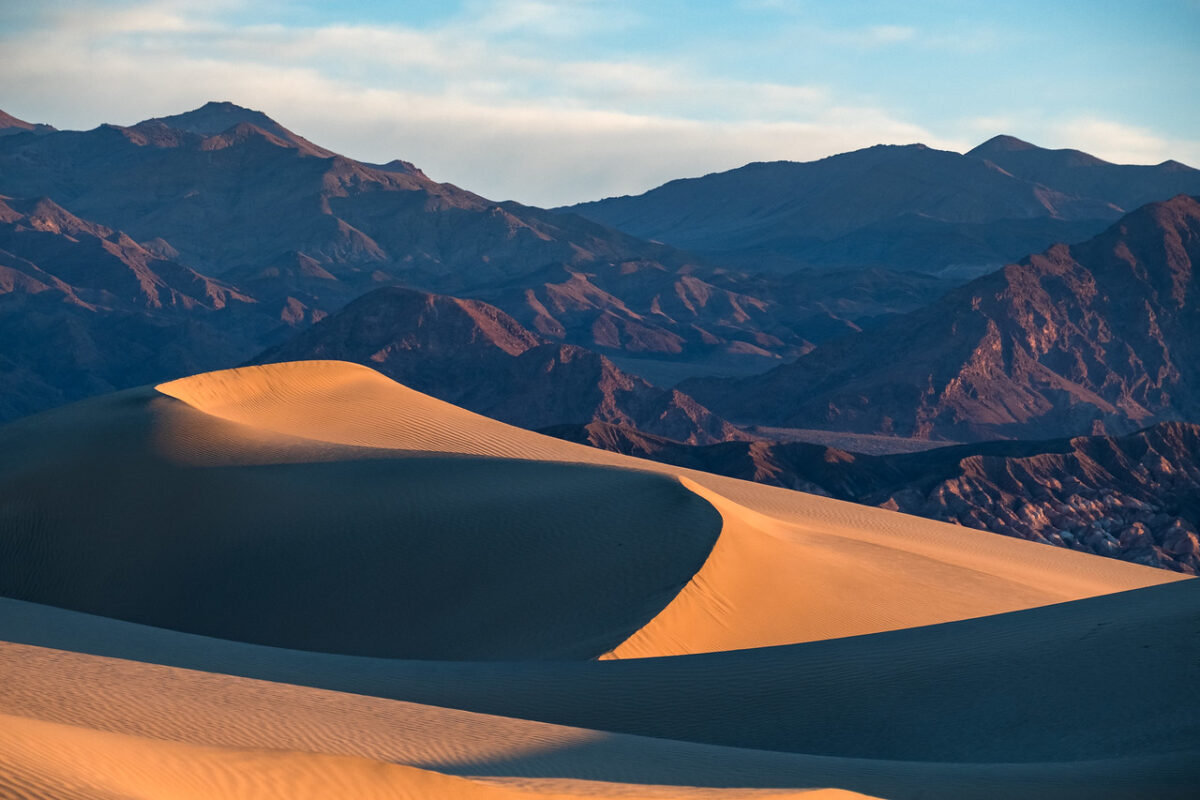 Sunlit sand dunes in Death Valley National Park curve across the foreground, their warm golden tones contrasting with deep shadows and layered mountain ranges beyond.