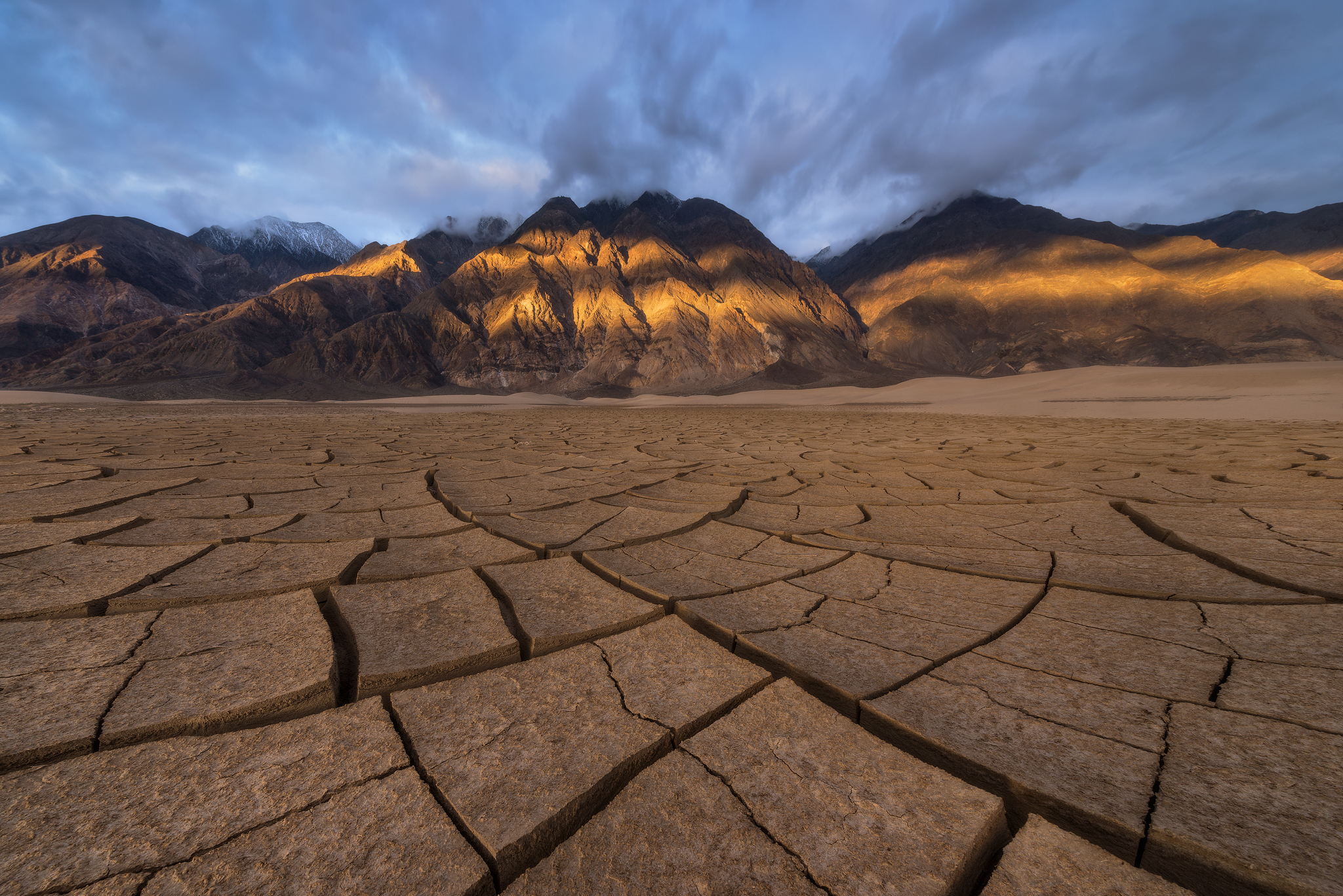 Large cracked mud polygons on a Death Valley playa create leading lines towards mountains illuminated by dappled sunlight beneath dramatic clouds.