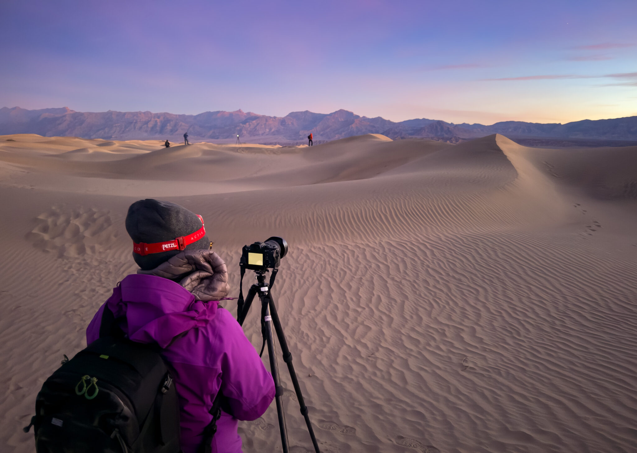 A photographer stands beside a tripod-mounted camera among sweeping sand dunes at dawn, with soft pastel light illuminating the desert landscape and distant mountains.