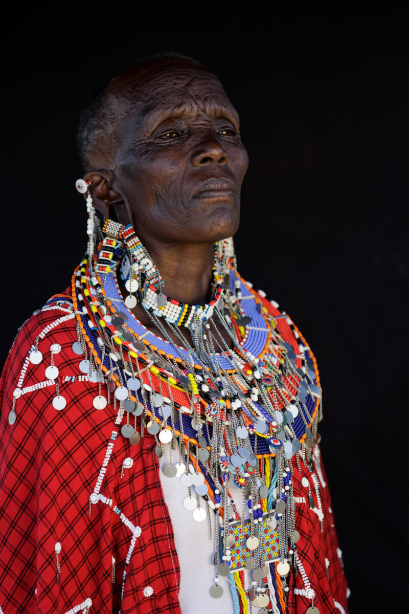 Maasai woman wearing traditional beaded jewellery and red clothing, photographed against a dark background in Kenya.