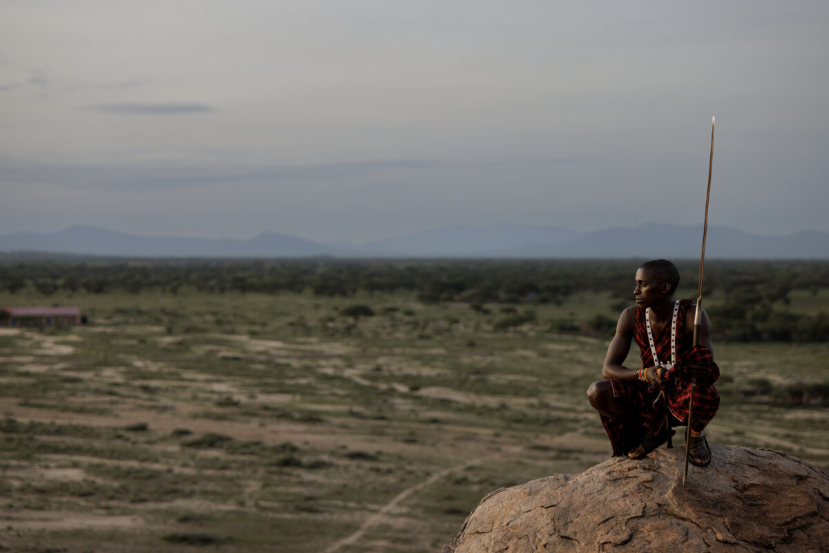 Young Maasai warrior overlooking the Kenyan landscape while standing on a rocky outcrop with a traditional spear.