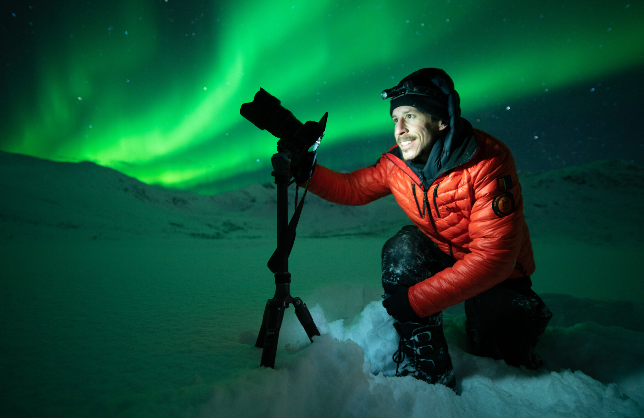 A photographer kneels in deep snow beneath a vivid green aurora borealis in northern Norway, adjusting a camera mounted on a tripod while snowy mountains fade into the darkness behind him.
