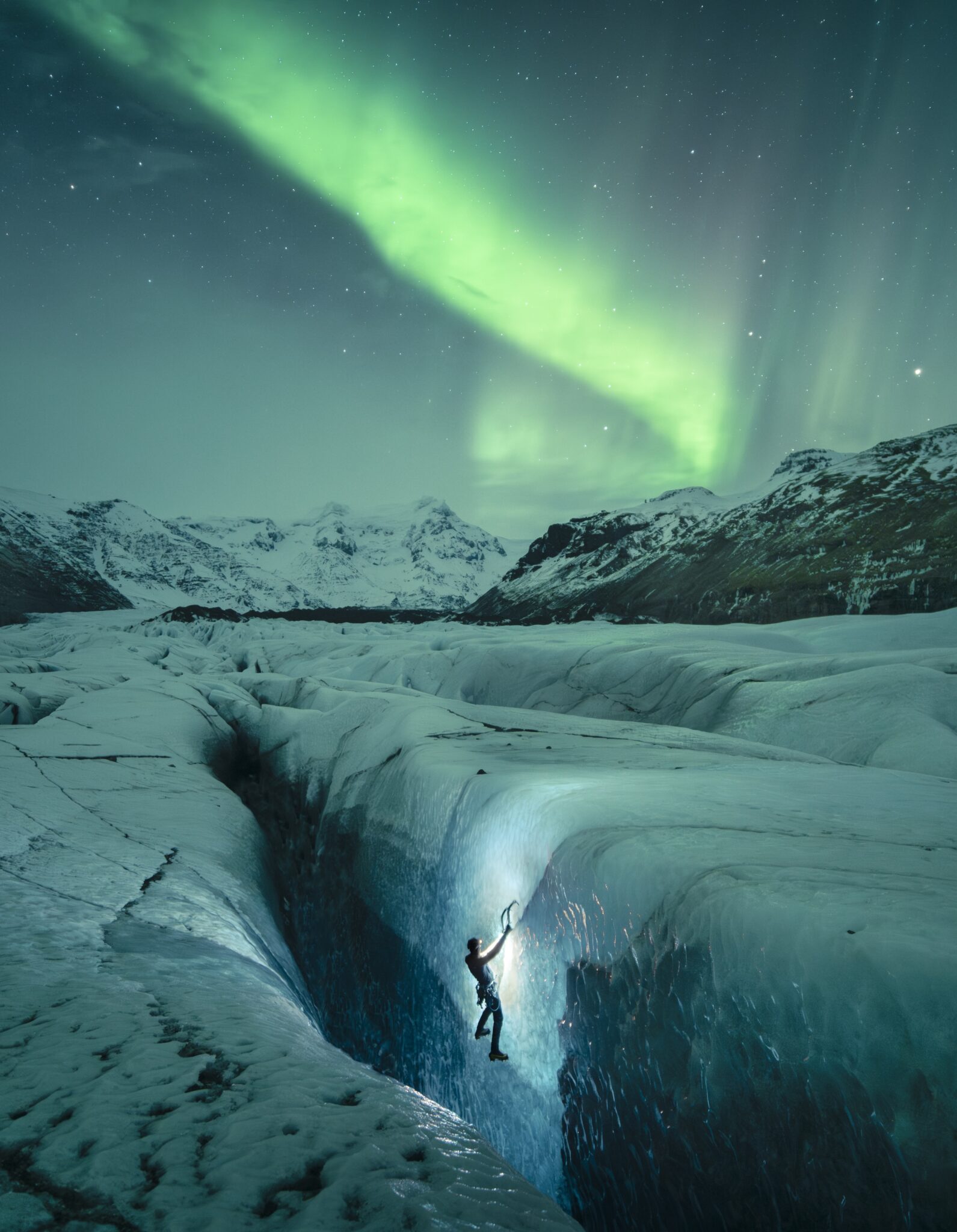 Ice climber descending into a glacier crevasse beneath bright green Northern Lights and snow-covered mountains in Iceland.