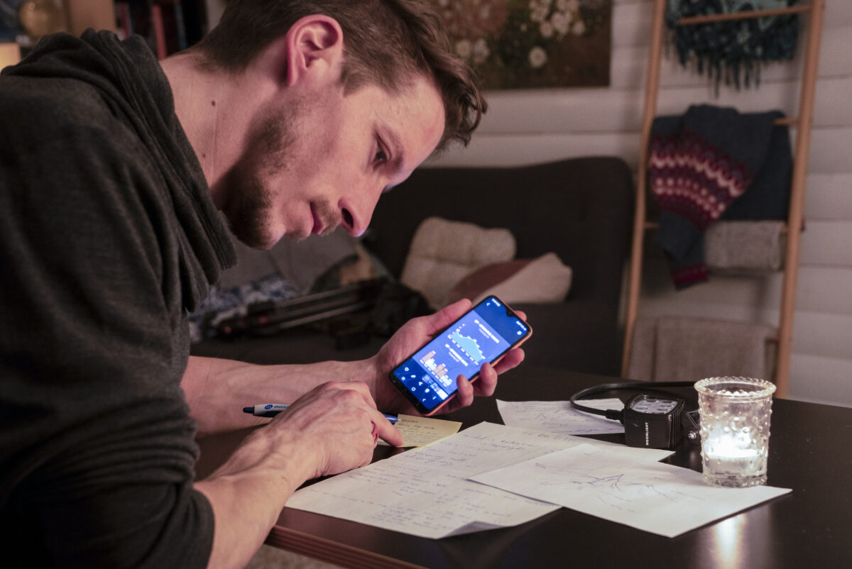 Photographer studying aurora forecast data on a smartphone while planning a Northern Lights shoot inside a warmly lit cabin.