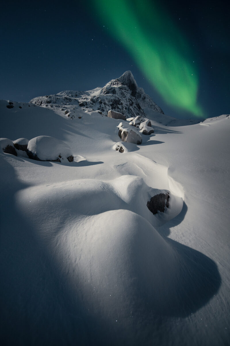 Moonlit snowdrifts leading toward a dramatic Arctic mountain peak beneath a ribbon of green Northern Lights.