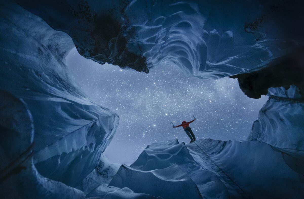 Person standing on the edge of a glacier crevasse beneath a star-filled sky, framed by sculpted blue ice walls and illuminated by the night.