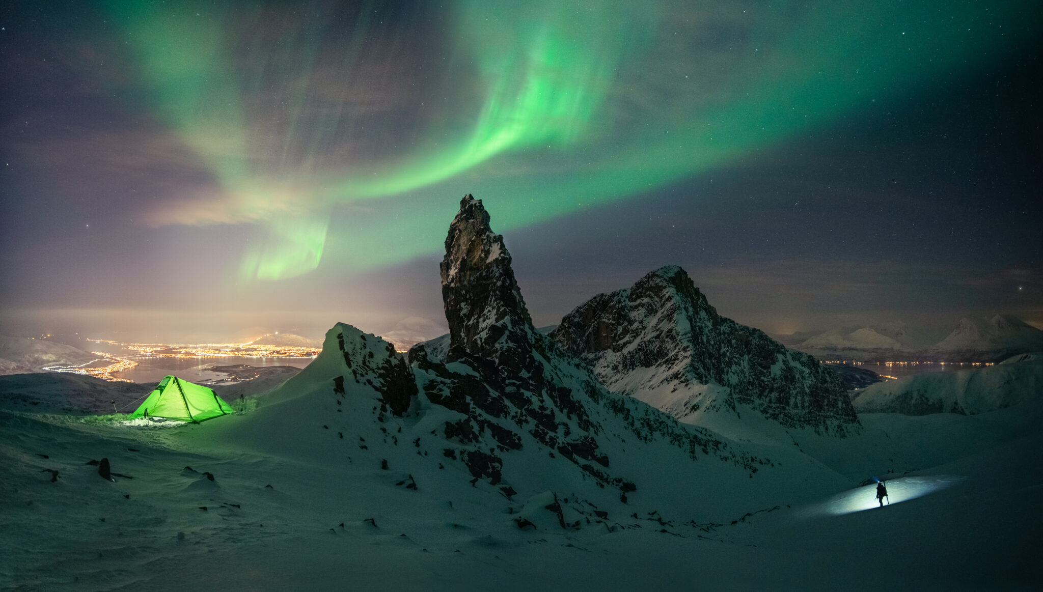 Northern Lights arching above a snowy mountain ridge, with a glowing green tent and lone hiker illuminated beneath the Arctic night sky.