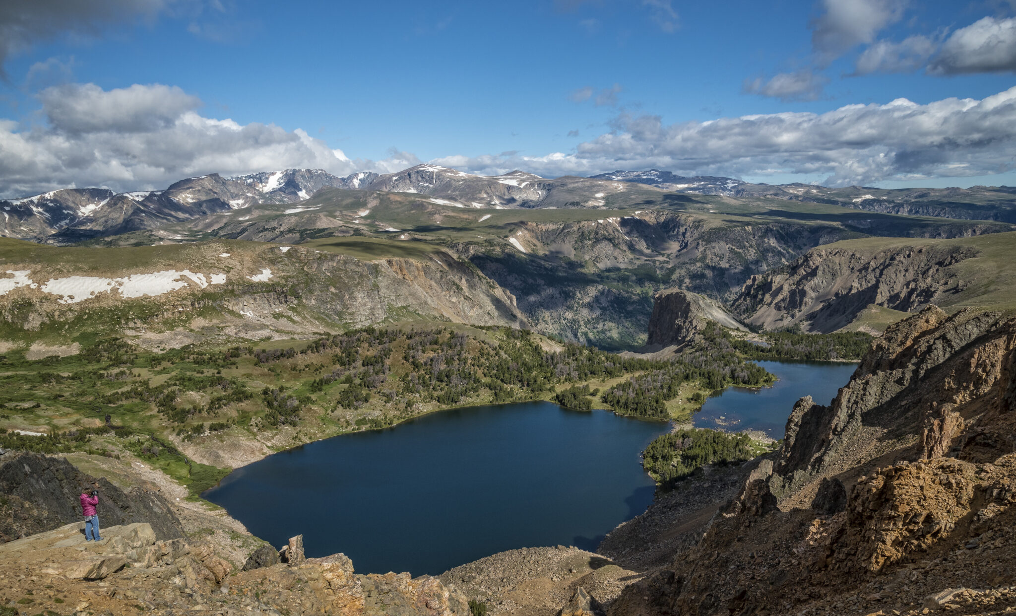 Panoramic view across the Beartooth Mountains, with a deep blue alpine lake nestled below rugged cliffs and high-elevation plateaus under a sky of scattered clouds. A lone photographer stands on a rocky overlook in the foreground.