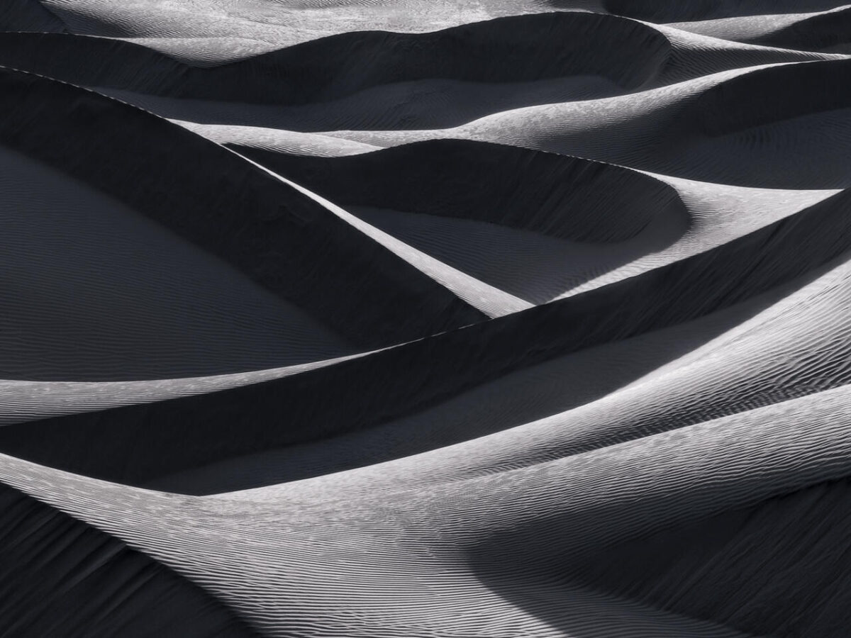 Abstract patterns of overlapping sand dunes in Death Valley National Park, where low-angle light creates sweeping curves, bright highlights and deep shadows across finely textured sand.