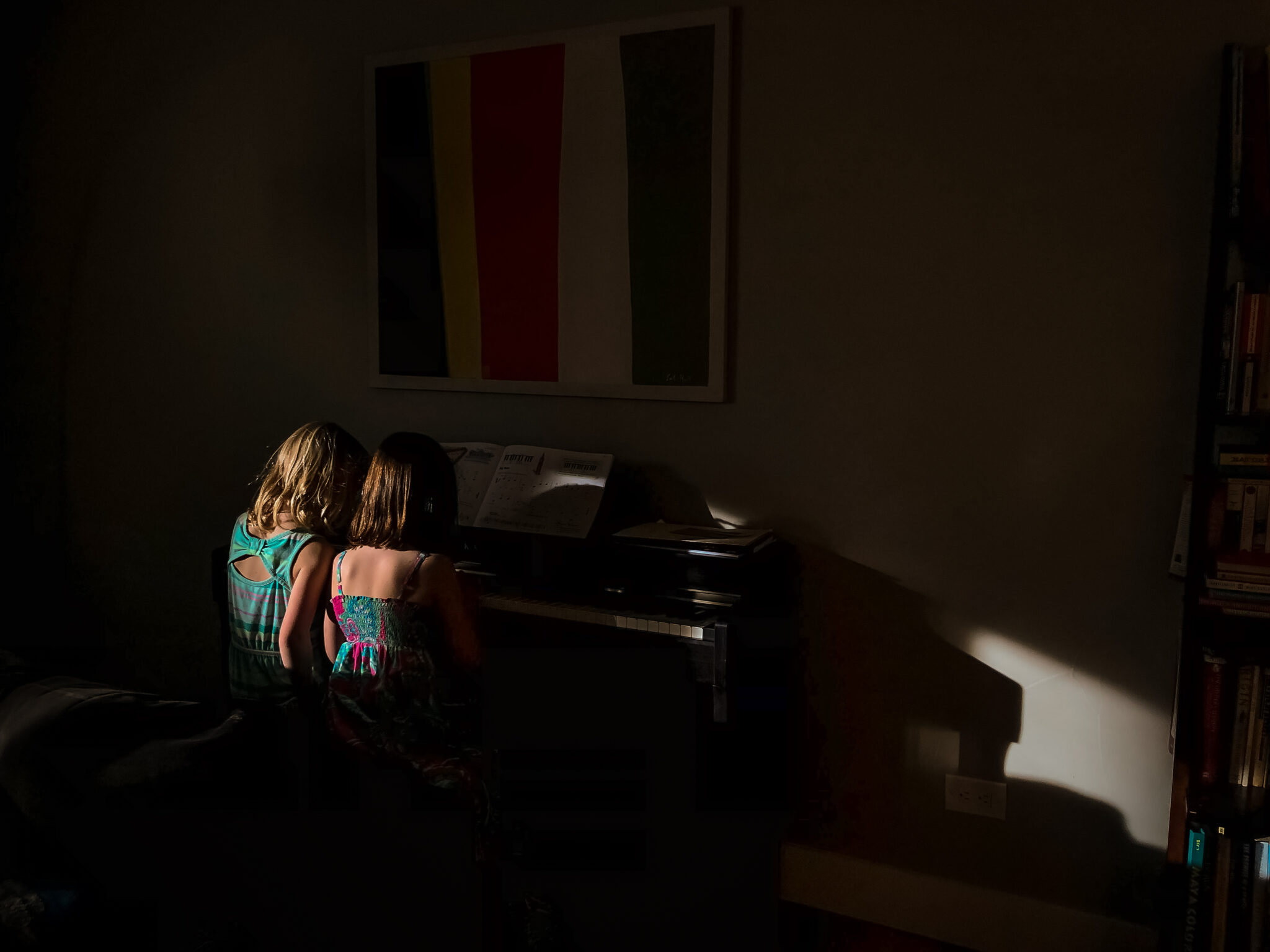 Two children sit at a piano in a darkened room, illuminated by a beam of warm afternoon sunlight as they play together.
