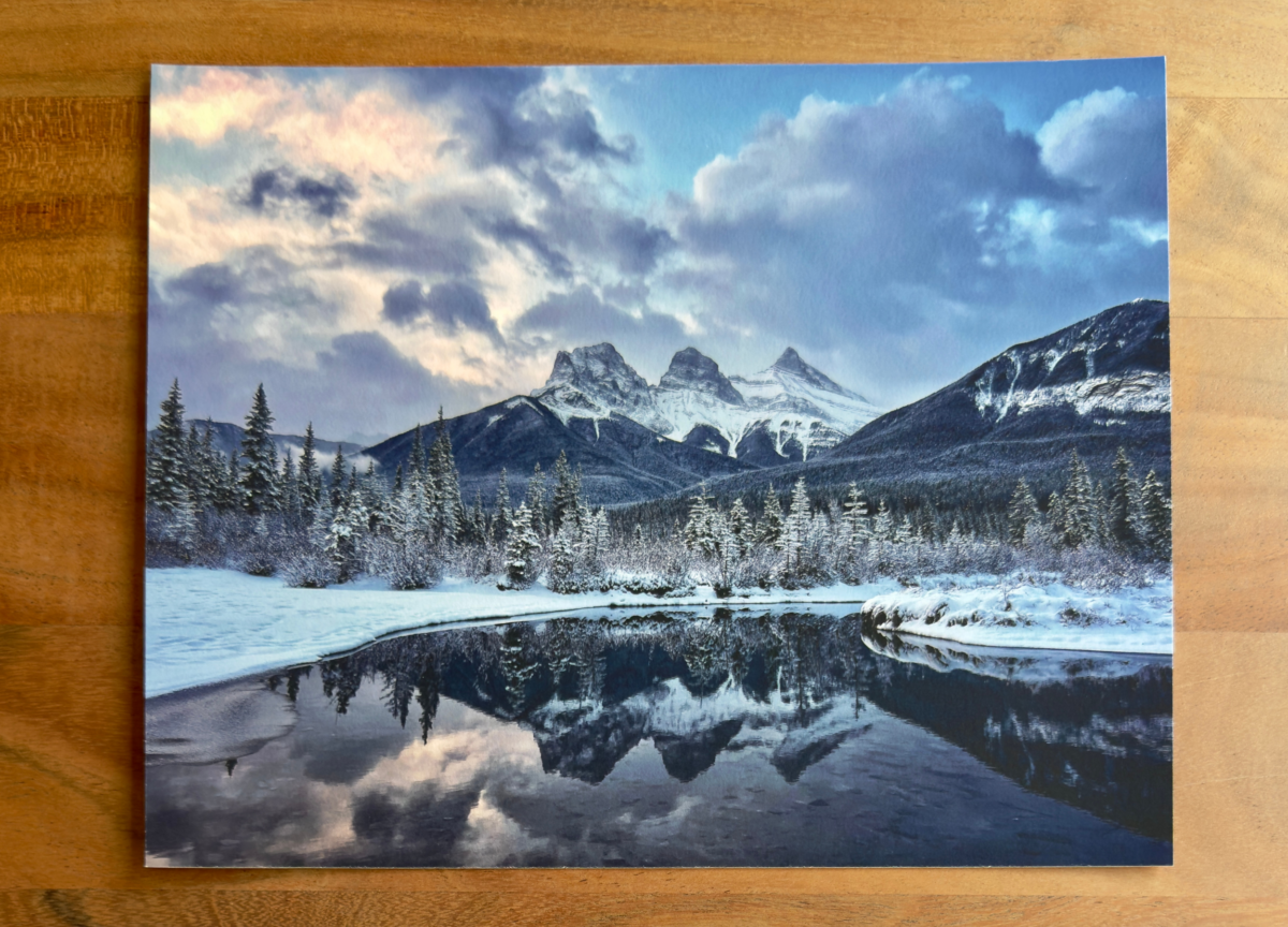 Two prints of a snow-covered mountain landscape with a reflective lake are displayed on a wooden table, comparing different print sizes.