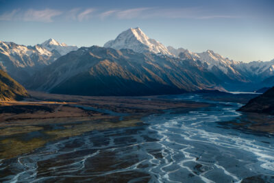 Panoramic view of Aoraki / Mount Cook rising above the braided riverbeds of the Tasman Valley in New Zealand's South Island. Snow-capped peaks glow in warm early morning light beneath a clear blue sky, while silver glacial streams wind across the expansive valley floor toward the mountains.
