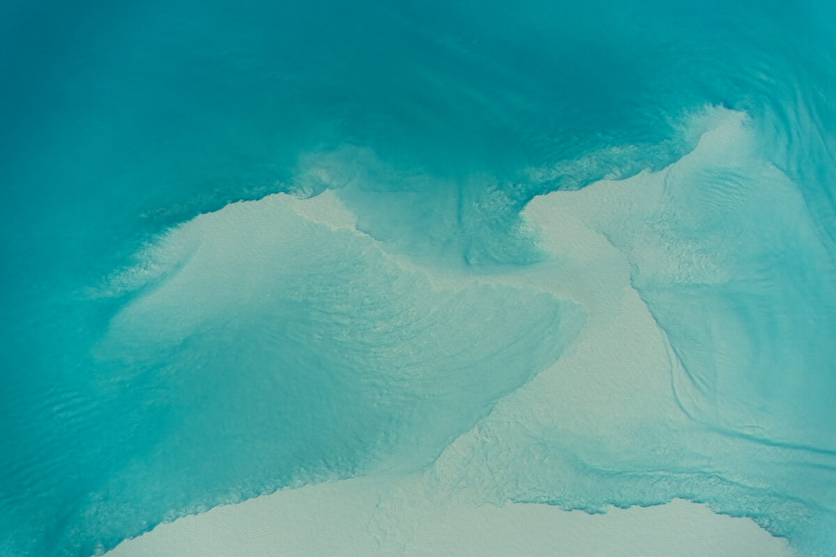 Abstract aerial view of turquoise glacial water flowing over pale sandbars, creating swirling patterns and soft gradients of blue and white. The natural shapes resemble flowing brushstrokes, highlighting the textures and movement of the braided river.