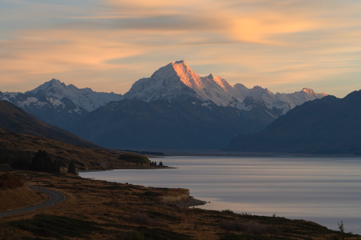 Aoraki / Mount Cook catches the first light of sunrise, its snow-covered summit glowing pink above the calm waters of Lake Pukaki. Soft pastel clouds stretch across the sky while the winding lakeside road and golden tussock-covered hills frame the peaceful South Island landscape.