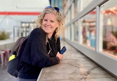iPhone photographer Kelley Dallas pauses between shoots outside a modern building, smiling while holding her phone.