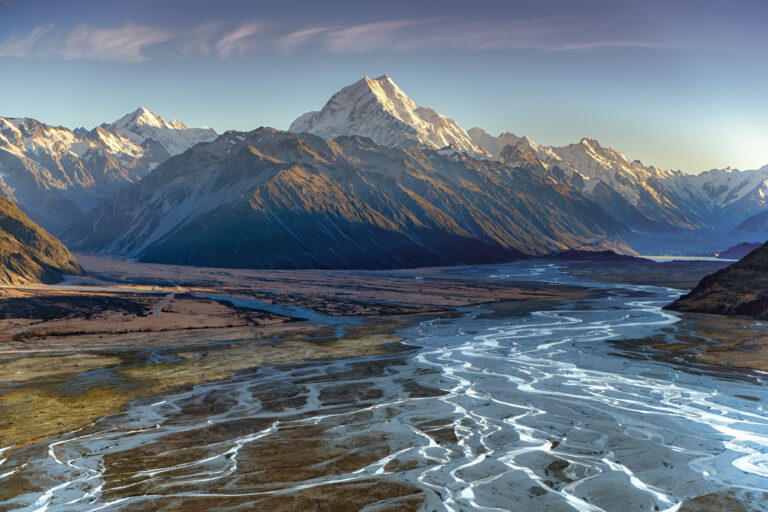 Panoramic view of Aoraki / Mount Cook rising above the braided riverbeds of the Tasman Valley in New Zealand's South Island. Snow-capped peaks glow in warm early morning light beneath a clear blue sky, while silver glacial streams wind across the expansive valley floor toward the mountains.