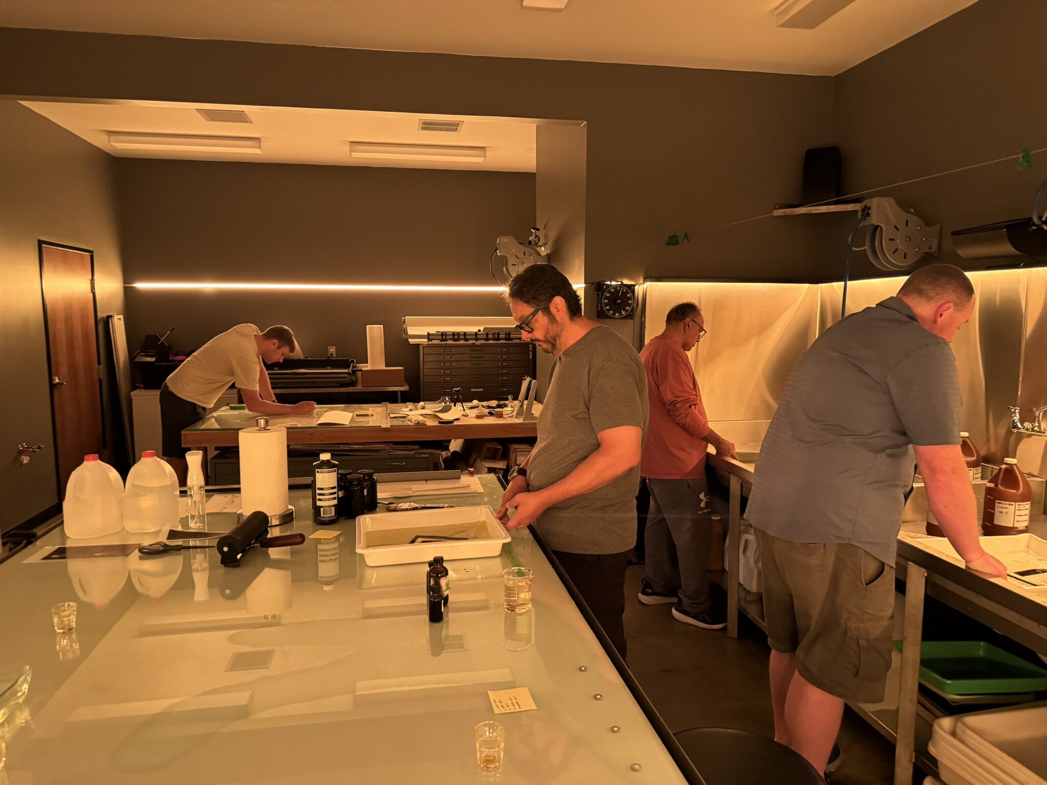 Participants work on black-and-white prints in a professional darkroom during a hands-on printing workshop. Several photographers prepare paper, develop prints in chemical trays, and make adjustments at workstations, with safelights illuminating the room and printing equipment arranged throughout the space.