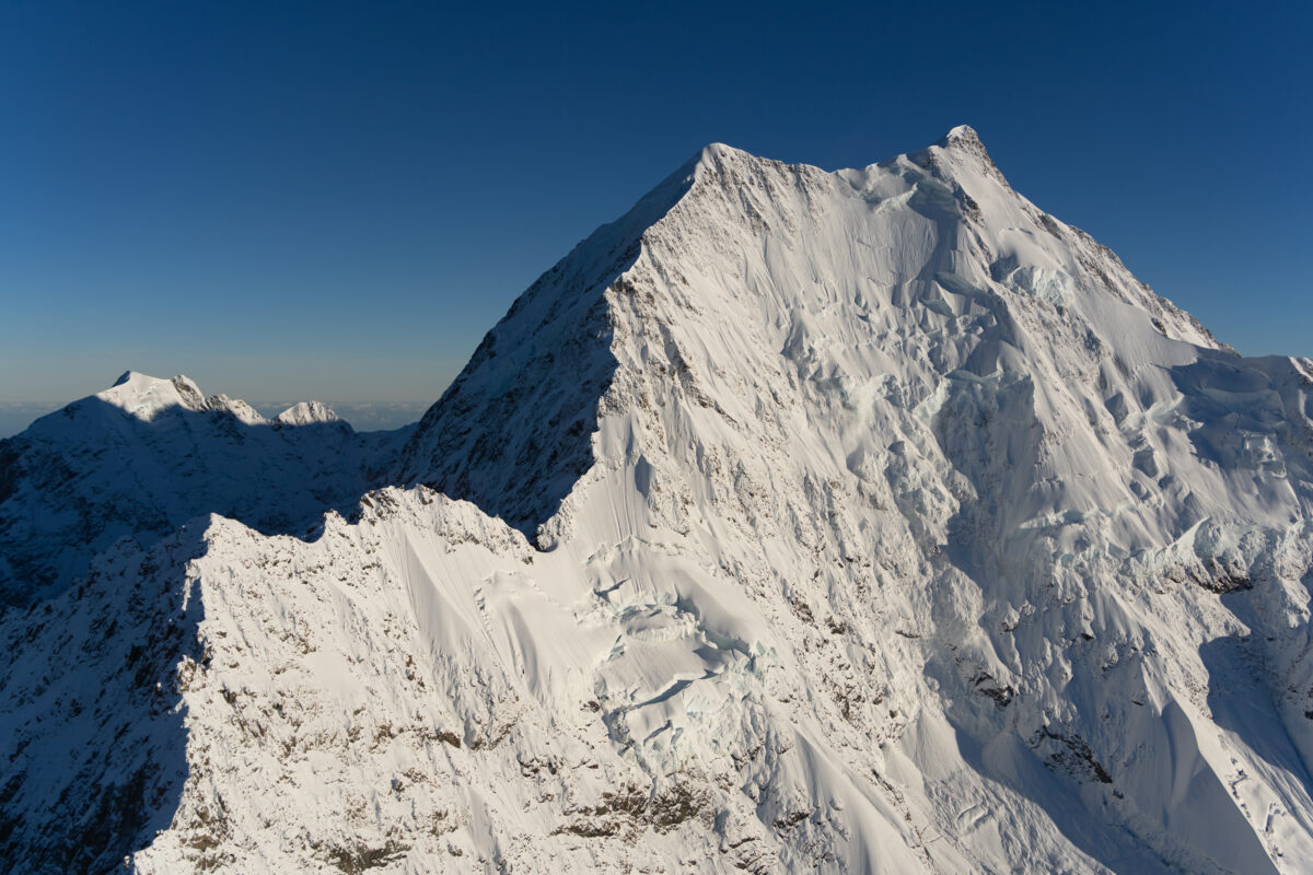 Close-up aerial view of a snow-covered peak in New Zealand's Southern Alps, revealing steep ridges, wind-sculpted snow, and deep crevasses beneath a cloudless blue sky. Bright alpine light highlights the mountain's dramatic textures and sheer scale.