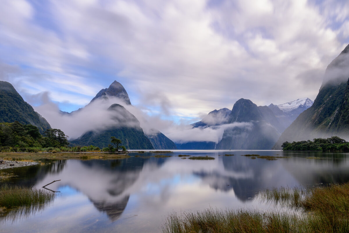 Long-exposure view of Milford Sound in Fiordland National Park, New Zealand, with mist swirling around Mitre Peak and surrounding mountains. Calm water reflects the dramatic peaks and fast-moving clouds, while low fog drifts across the fjord beneath a soft blue and grey sky.