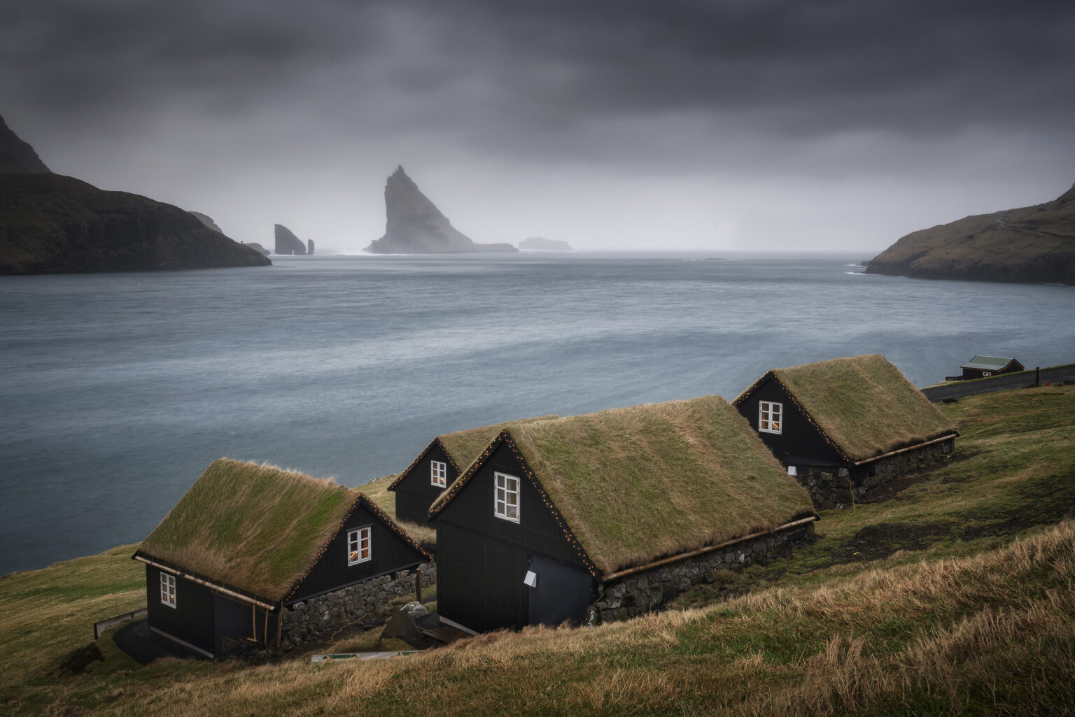 Traditional black wooden cottages with turf grass roofs on a grassy hill overlooking dramatic sea stacks under moody, overcast Faroe Islands skies.