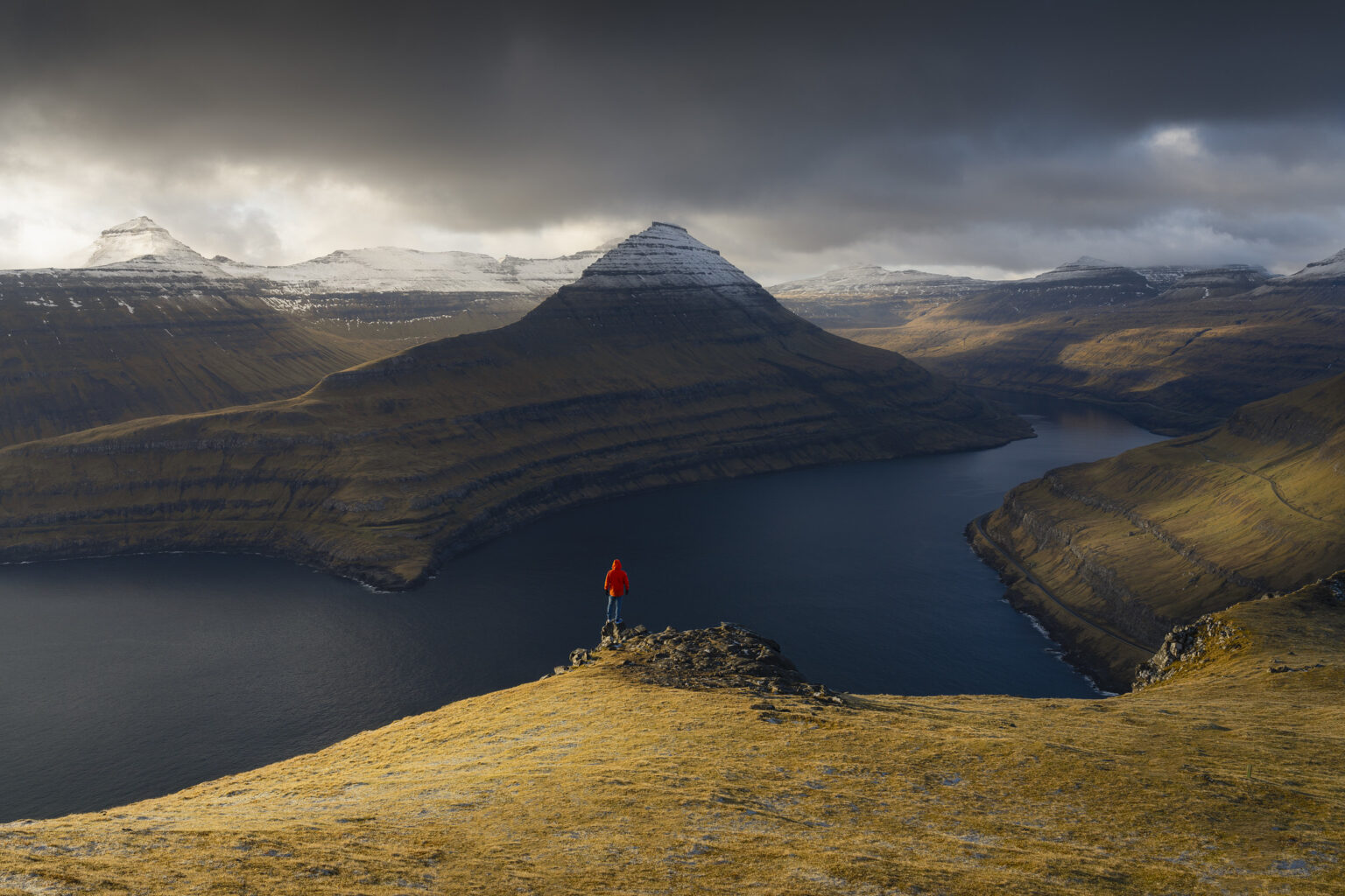 A person in a bright red jacket standing on a grassy cliff edge overlooking a deep fjord surrounded by snow-capped mountains.