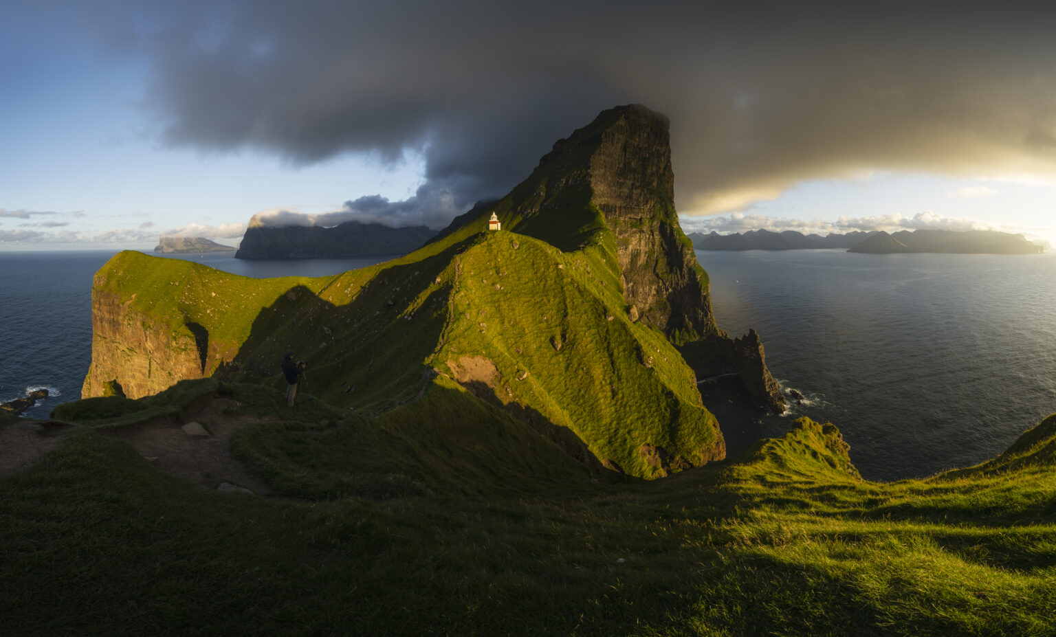 Sunlit green ridge leading to Kallur Lighthouse atop steep ocean cliffs in the Faroe Islands.