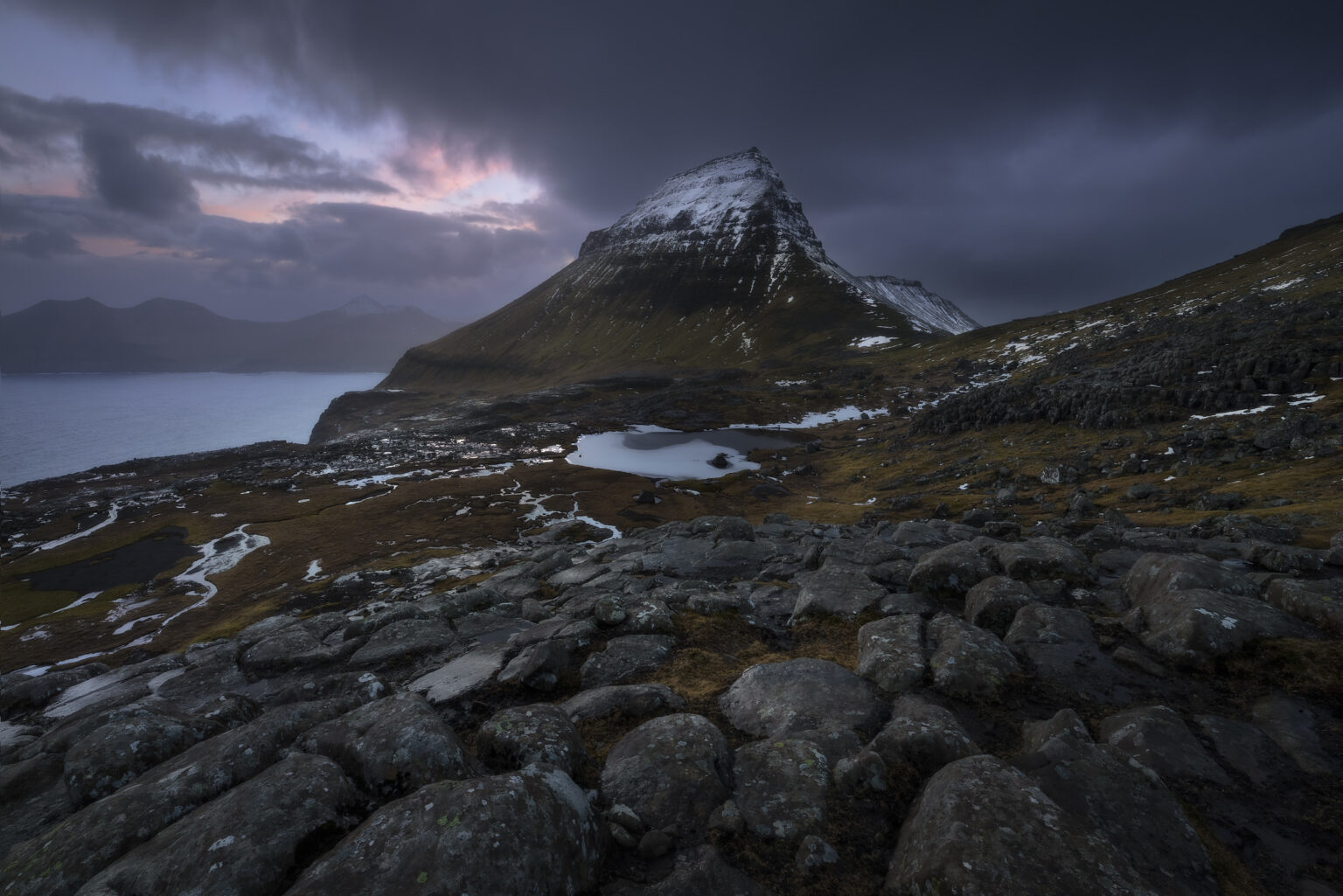 A rocky foreground leading to a snow-capped mountain peak and small glacial pool beneath a dark, dramatic sunrise sky in the Faroe Islands.