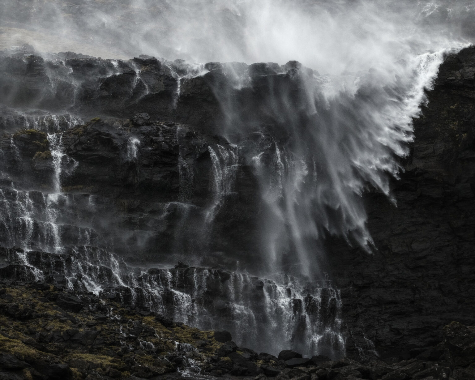 Strong winds blowing waterfall spray back up dark, rocky cliff faces in a dramatic Faroese landscape.