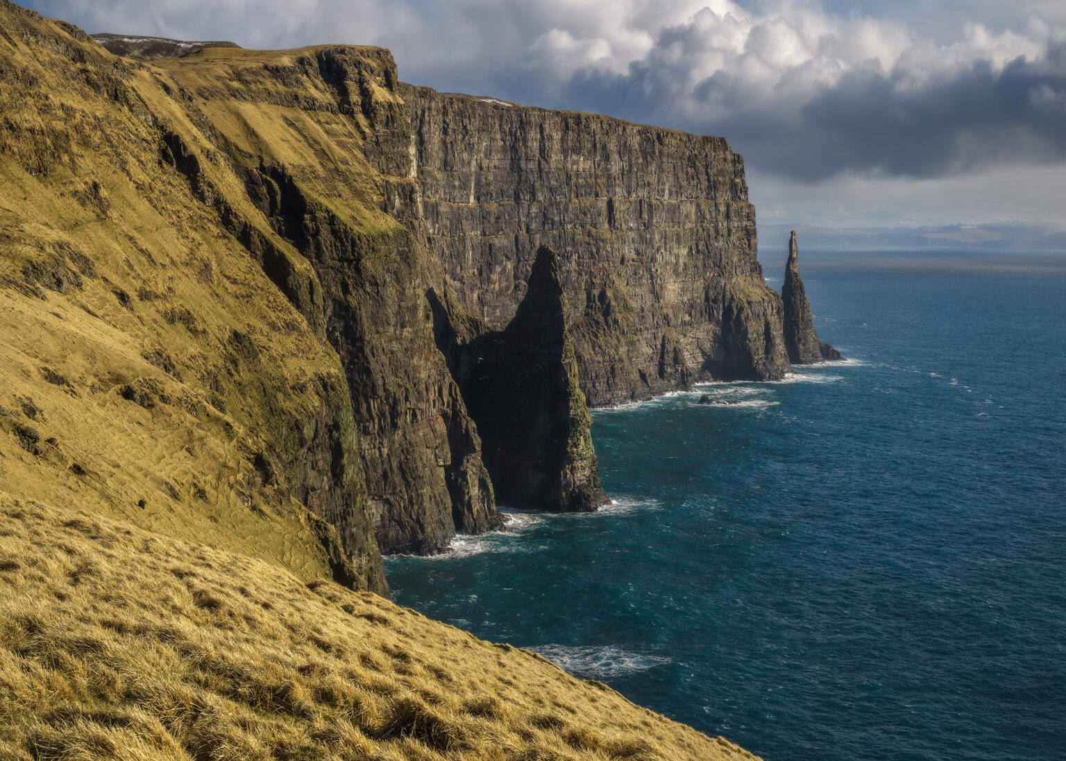 A dramatic wide shot of a grassy, steep slope leading down to massive vertical rock cliffs and sea stacks jutting out of deep blue ocean water. Bright sunlight illuminates the yellowish-green grass on the mountain side, contrasting with the shadowed rock face and cloud-filled sky.