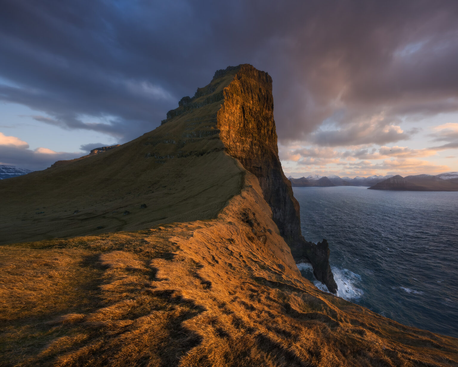 A panoramic view of a massive, pointed grassy ridge and cliff illuminated by warm, golden sunset light. The steep headland drops sharply into a dark blue sea on the right, with distant island peaks visible on the horizon under a moody, cloud-covered sky.