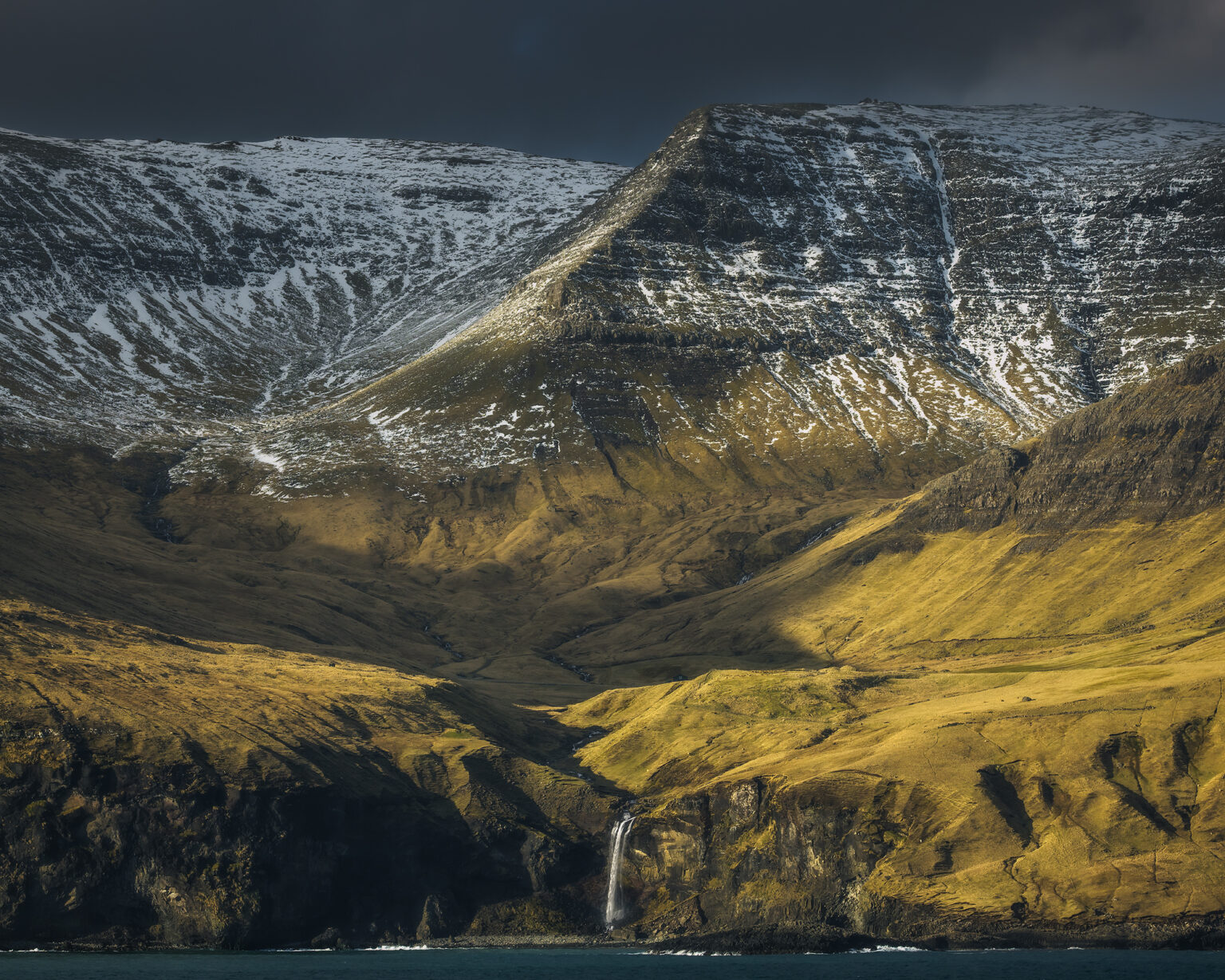 A dramatic wide shot of a grassy, steep slope leading down to massive vertical rock cliffs and sea stacks jutting out of deep blue ocean water. Bright sunlight illuminates the yellowish-green grass on the mountain side, contrasting with the shadowed rock face and cloud-filled sky.