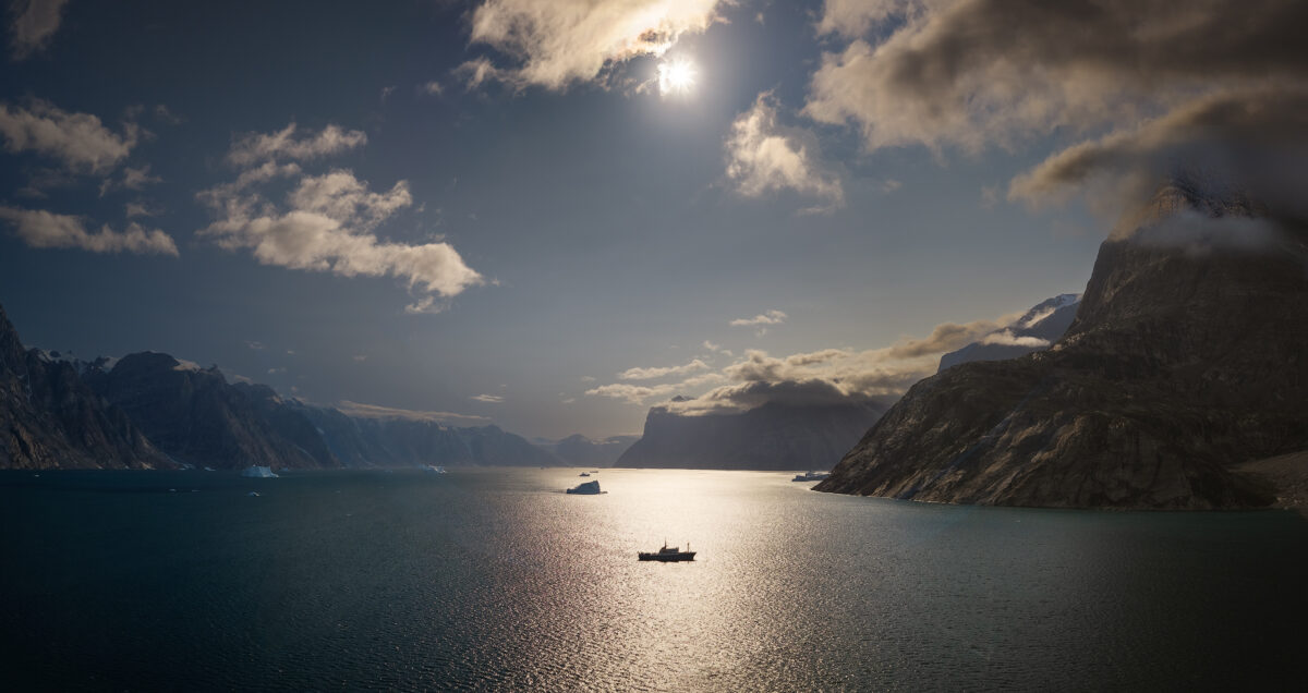 A panoramic view of a vessel navigating a wide Arctic fjord, flanked by massive cloud-capped mountains with sunlight reflecting off the water.