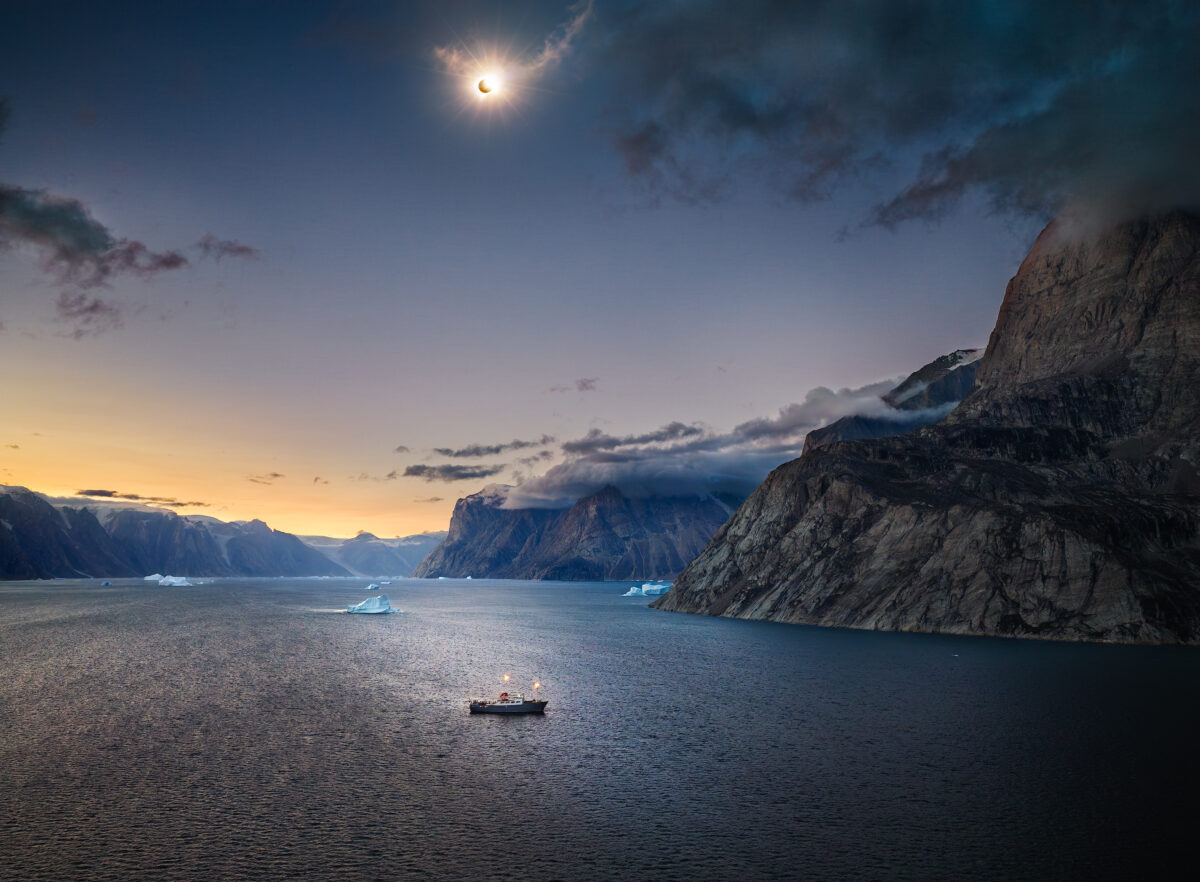 A small vessel floats in a tranquil Arctic fjord surrounded by towering, cloud-draped mountain cliffs, with a glowing solar eclipse illuminating the twilight sky above.