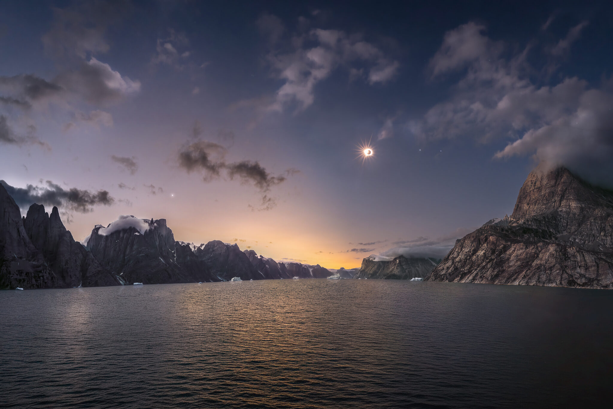 A wide panoramic view of a deep Arctic fjord flanked by towering, dark granite cliffs, with a total solar eclipse glowing in the darkening sky above.