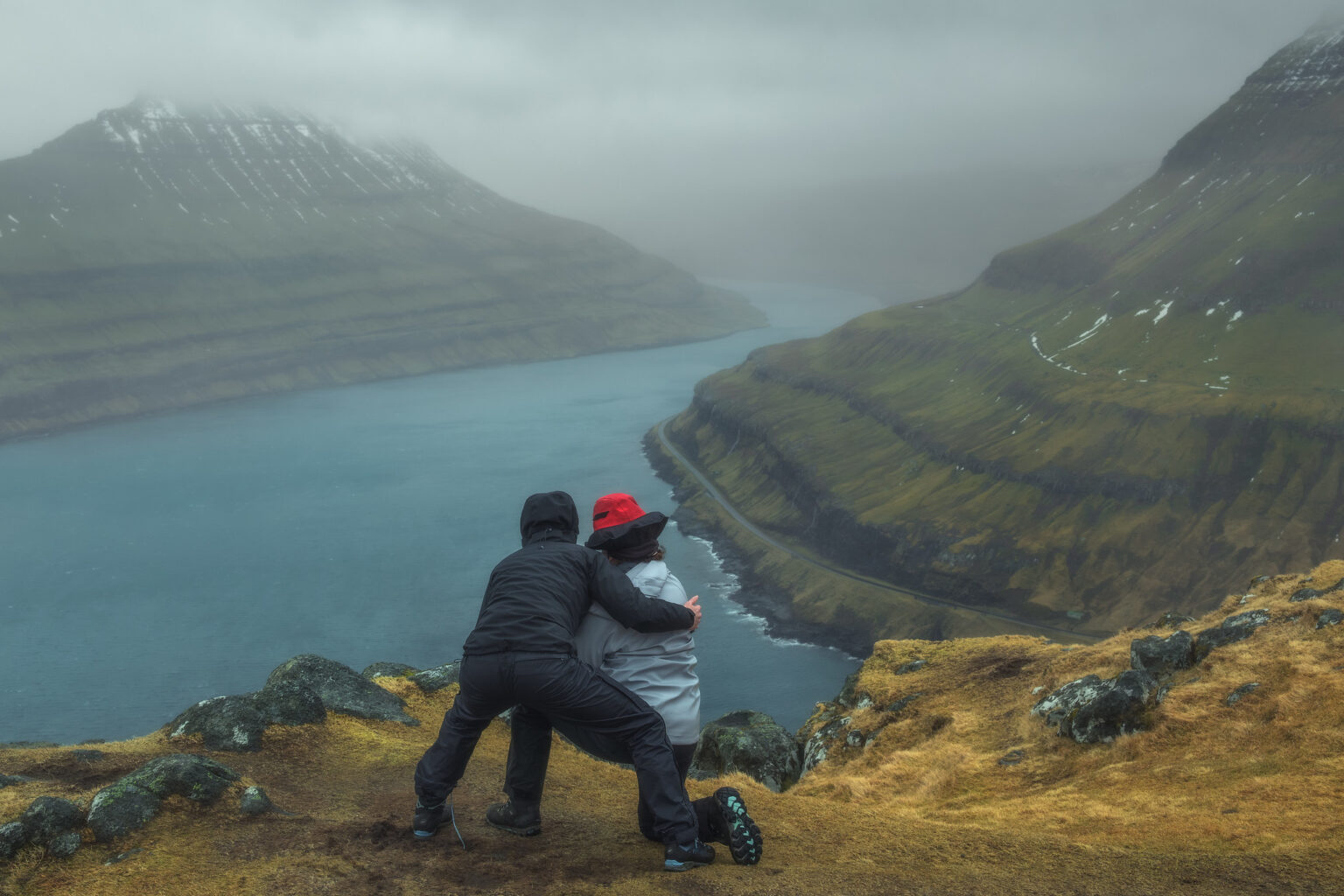 Two photographers wearing winter gear embracing on a high cliff top, looking out over a foggy fjord in the Faroe Islands.