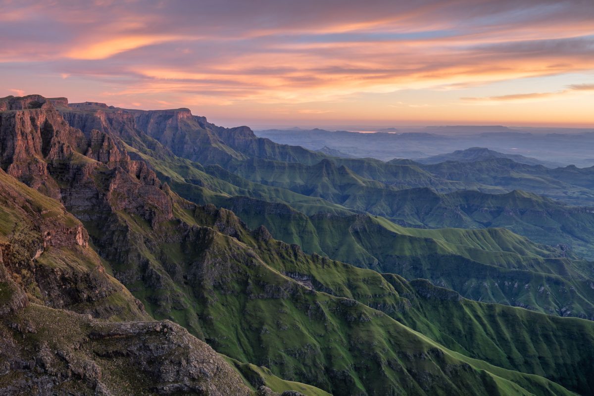 A vast panorama of the Drakensberg mountain range featuring dramatic green ridge lines glowing under a soft pink and orange sunset sky.