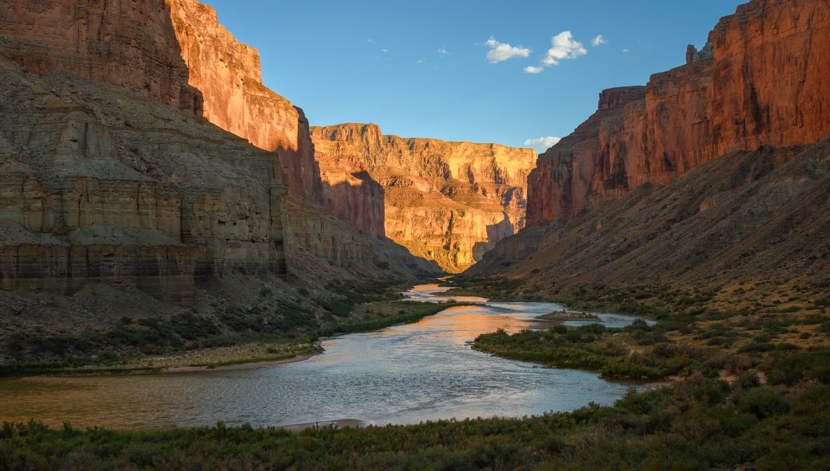 The Colorado River winding through the steep, red-rock cliffs of the Grand Canyon, illuminated by warm evening light.