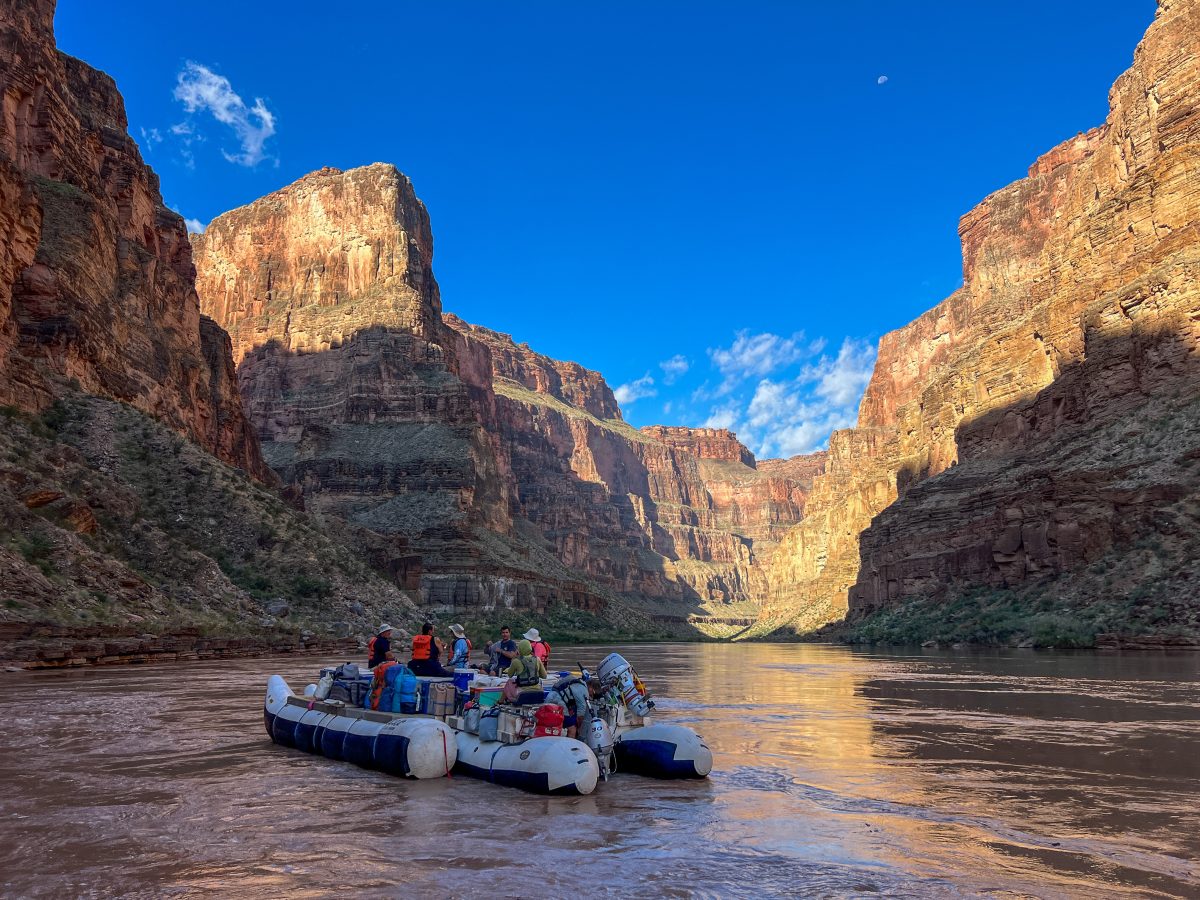 Rafts carrying a group of people along the Colorado River between towering cliffs in the Grand Canyon.