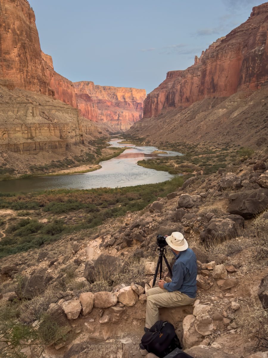A photographer wearing a wide-brimmed hat sits beside a tripod on a rocky hillside, photographing the winding Colorado River and towering red canyon cliffs in warm evening light.