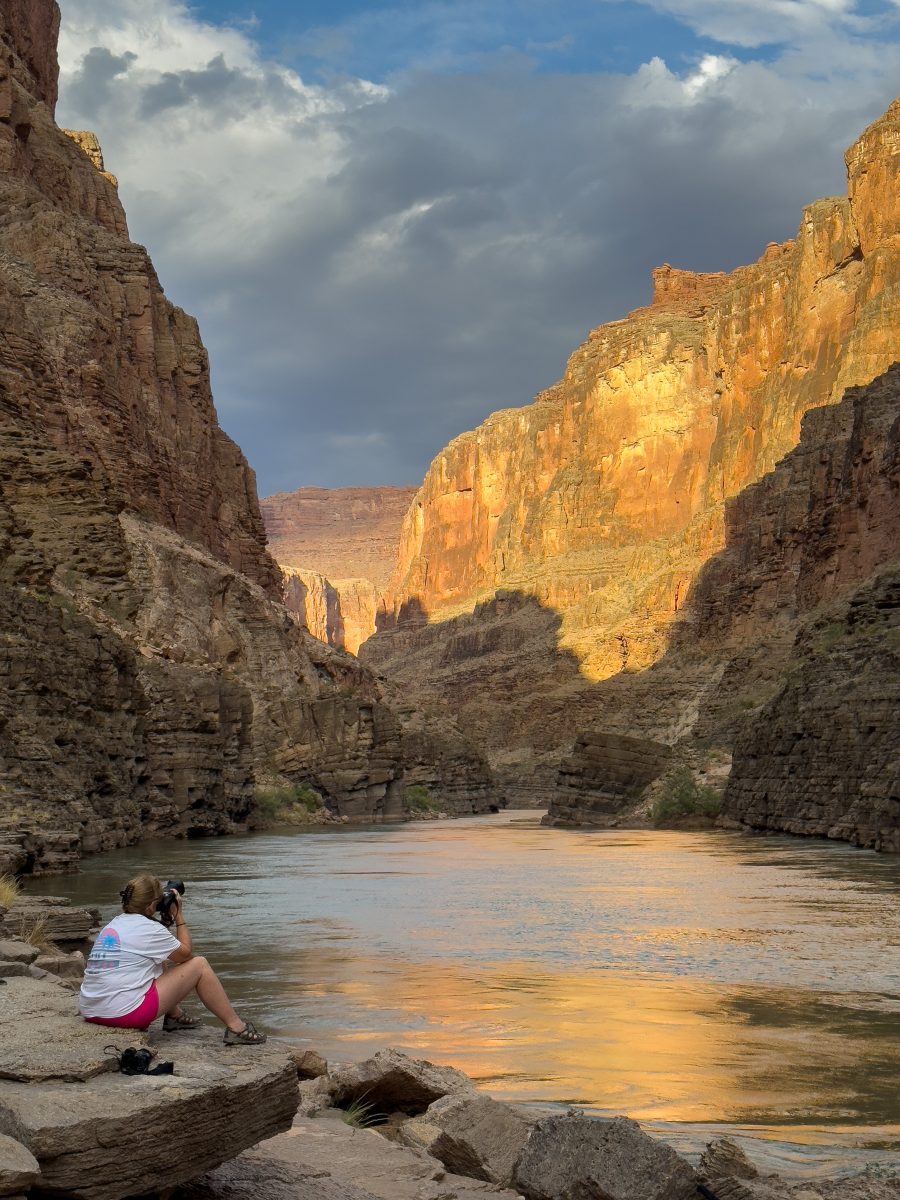 A photographer sits on rocks beside the Colorado River, photographing golden sunlight illuminating the towering canyon walls beneath a dramatic cloudy sky.