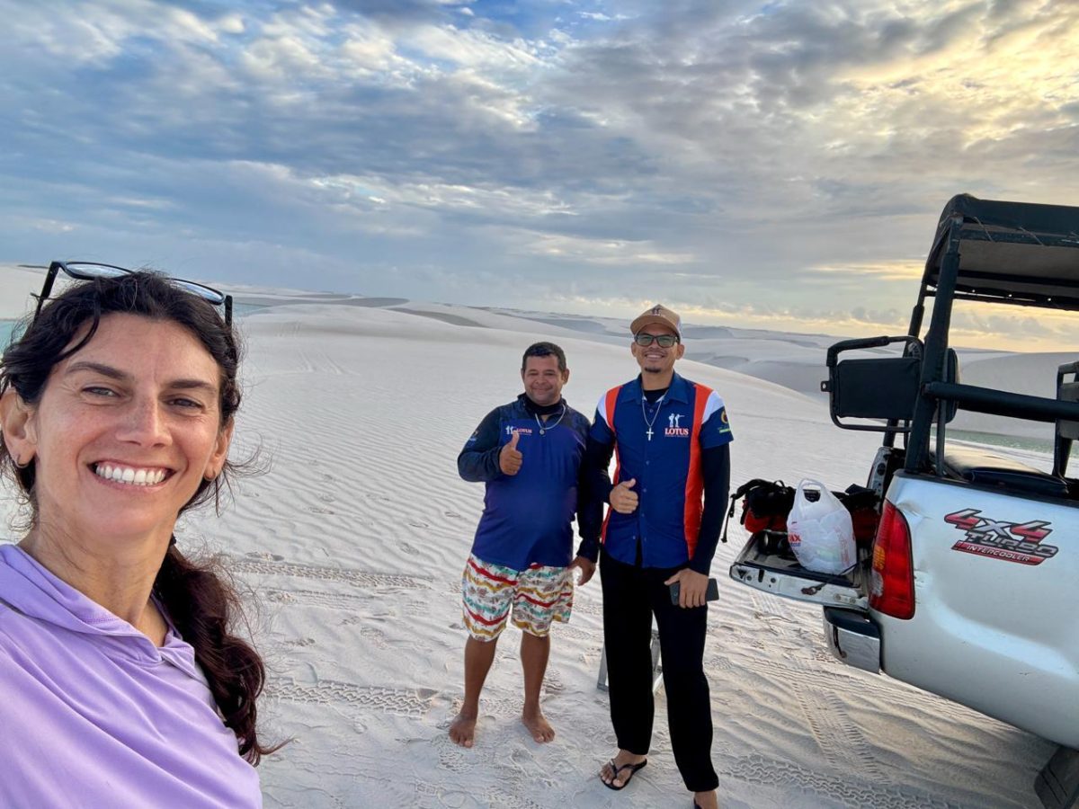 Three people standing beside a 4x4 vehicle on expansive white sand dunes, smiling for a selfie beneath a partly cloudy sky.
