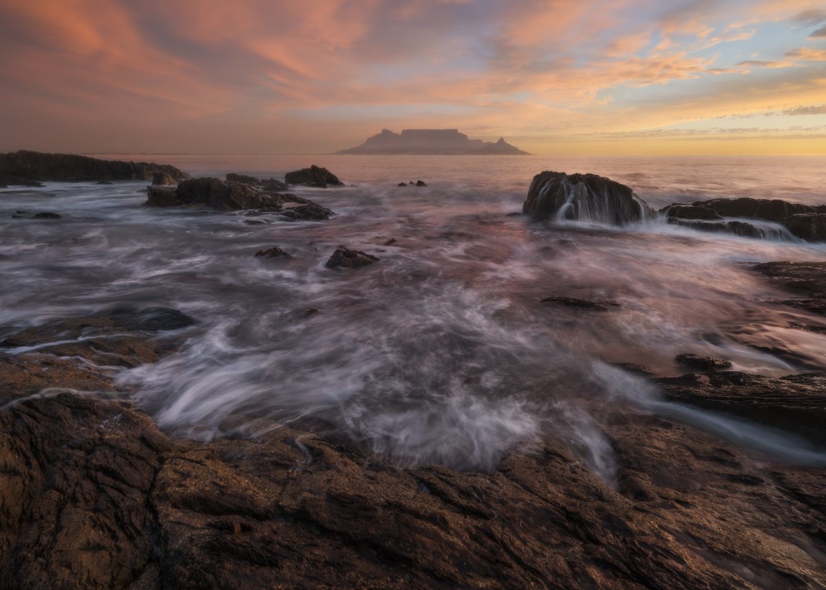 Long-exposure seascape at sunset, with waves flowing around dark rocky outcrops and Table Mountain silhouetted on the horizon beneath a warm pink and orange sky.