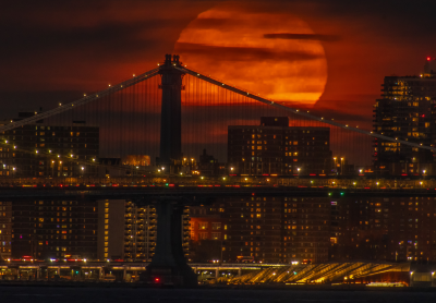 A huge orange moon rises behind the Manhattan Bridge, with its suspension cables silhouetted against the moon and illuminated Brooklyn buildings below.